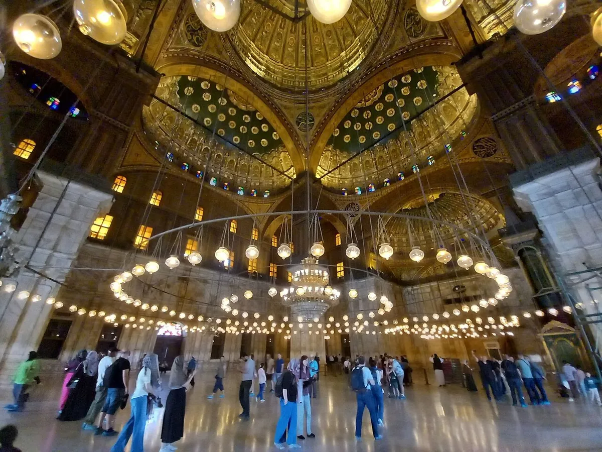 Visitors standing under the hanging lamps and painted domes inside the Muhammad Ali Mosque