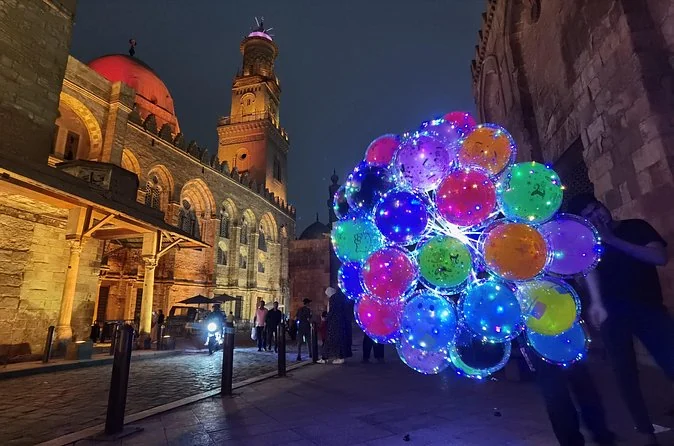Al-Muizz Street at night with lit facades, a minaret and a seller holding a bunch of glowing balloons